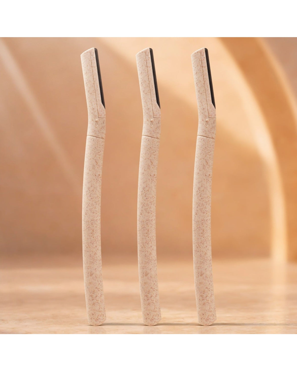 Three dermaplaning tools standing upright on a wooden surface against a warm beige background.