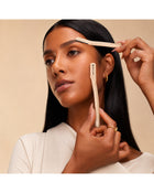 Woman using dermaplaning tools on her brow and cheek to shape brows and smooth facial hair.