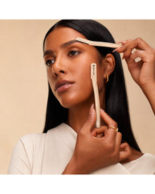 Woman using dermaplaning tools on her brow and cheek to shape brows and smooth facial hair.